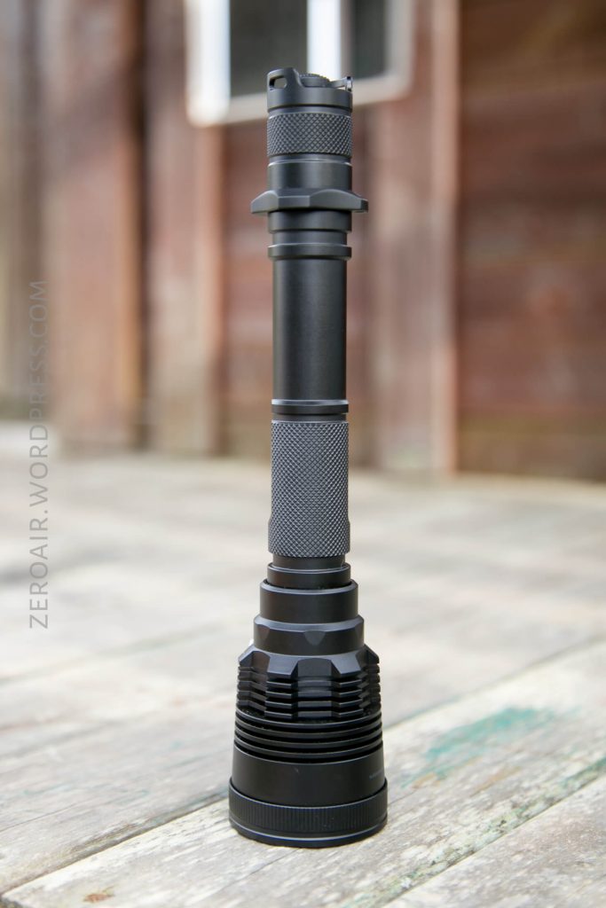 A black, cylindrical flashlight with a textured grip stands upright on a weathered wooden surface. A building with brown wooden panels and a window is blurred in the background.