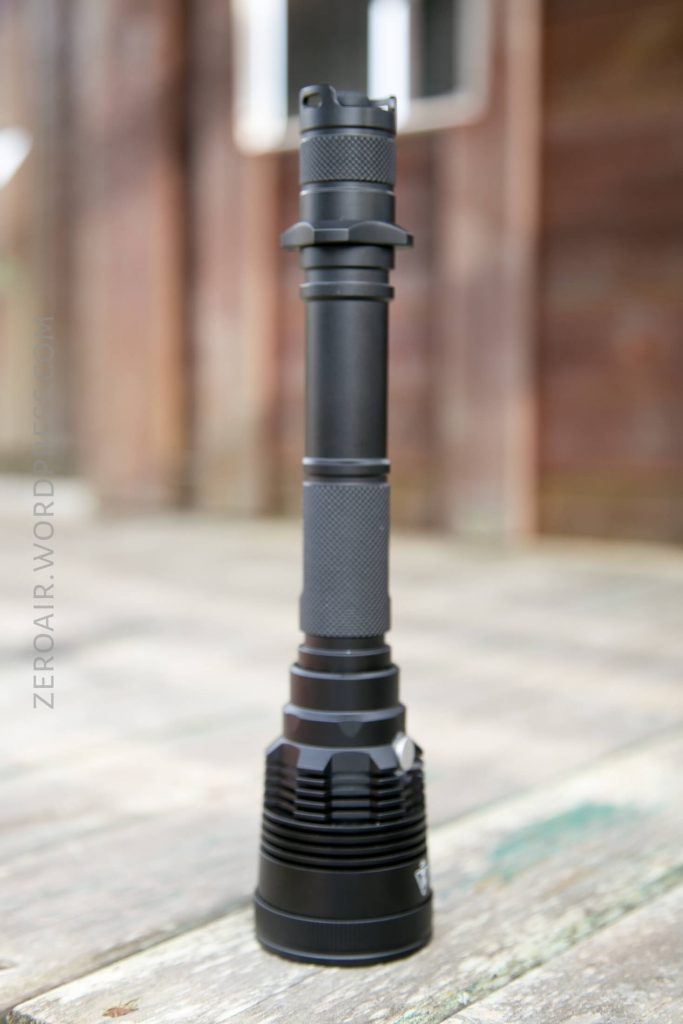 A black, heavy-duty flashlight stands upright on a wooden surface outdoors. The background is blurred, showing a wooden structure and a window.
