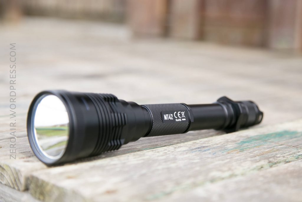 A black handheld flashlight with a wide lens lies on a weathered wooden surface. The flashlight has visible branding and safety markings on its side.