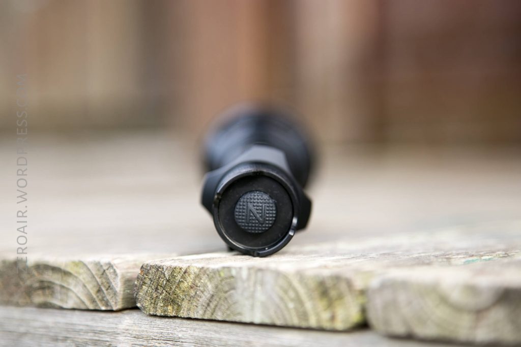 A black cylindrical object, possibly a flashlight, lies horizontally on a wooden surface. The focus is on the textured button at the end, featuring the letter N in the center. The background is blurred.