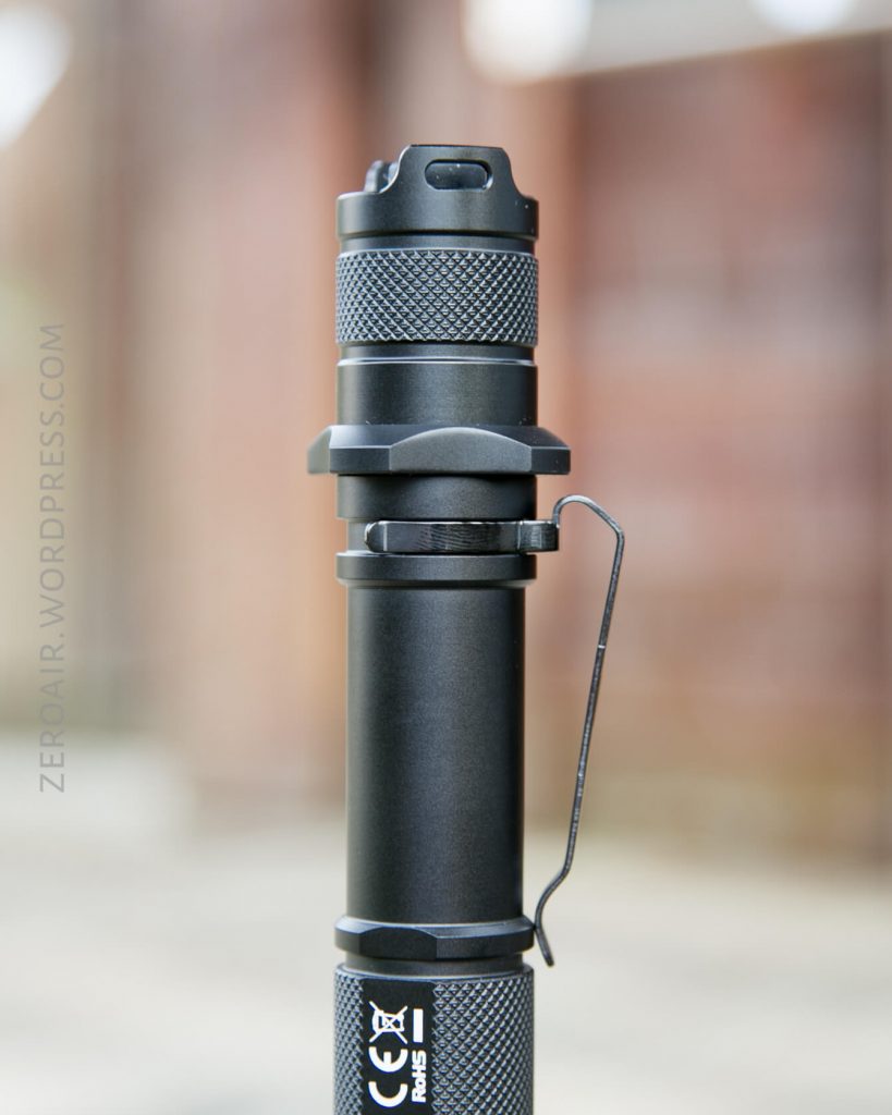A close-up of a black tactical flashlight with a textured grip, pocket clip, and visible branding. The background is blurred, focusing attention on the flashlight’s details.