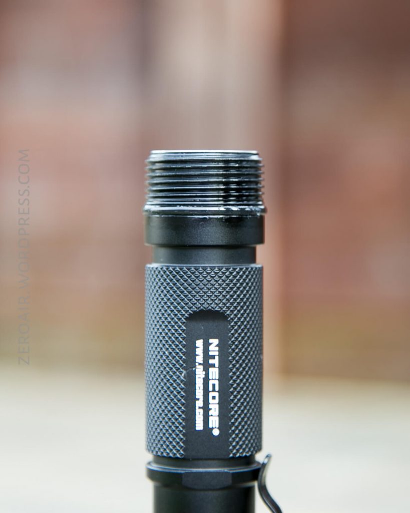 A close-up of a Nitecore flashlight with its head removed, showing the exposed screw threads on top of the textured metallic body. The background is blurred.
