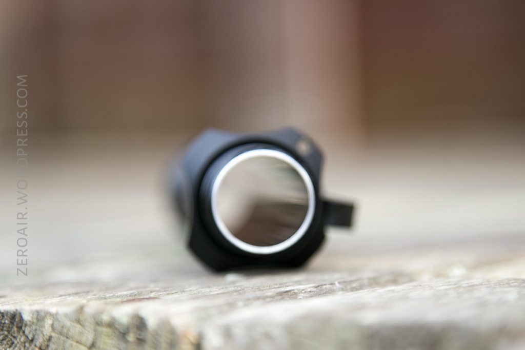 A close-up, front-facing view of a black cylindrical object resting on a wooden surface, with the background and object edges blurred due to shallow depth of field. The object appears to be metallic.