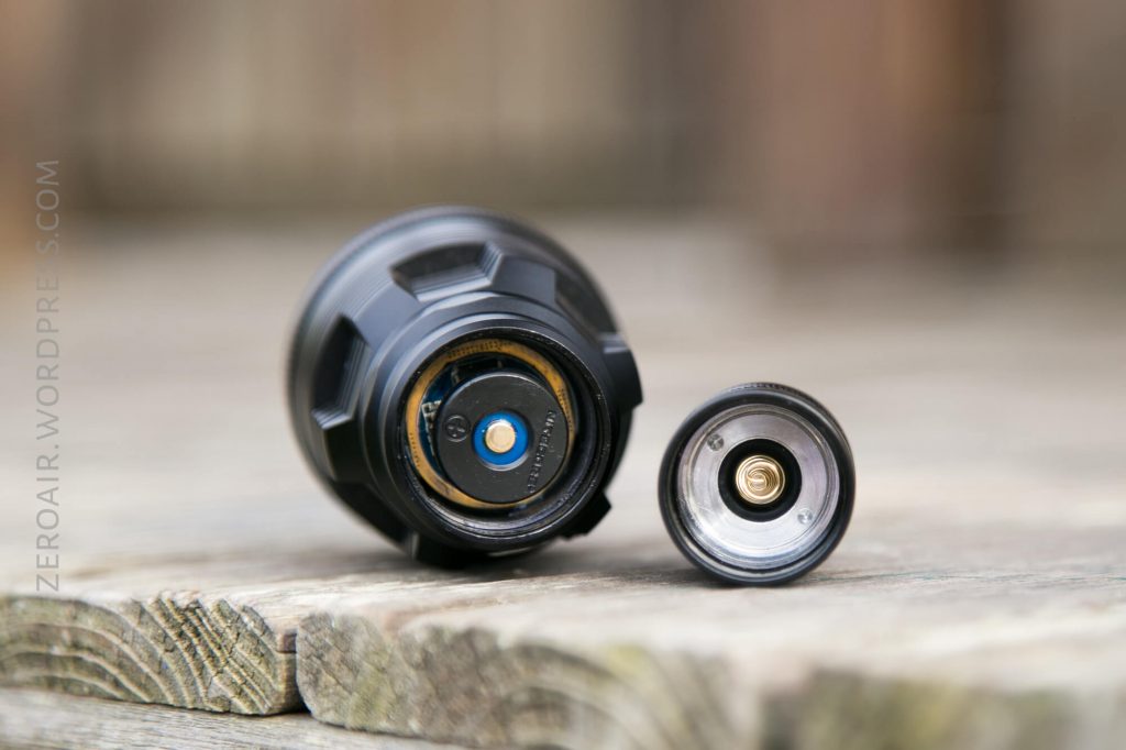 A close-up view of a disassembled flashlight and its battery compartment, placed on a wooden surface. The metal contacts and internal components are visible.
