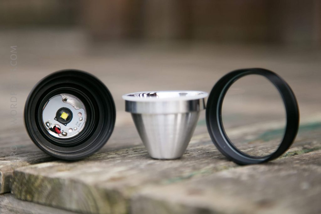 Three separate components of a flashlight, including the LED module, a reflective cone, and a black ring, are arranged side by side on a wooden surface.