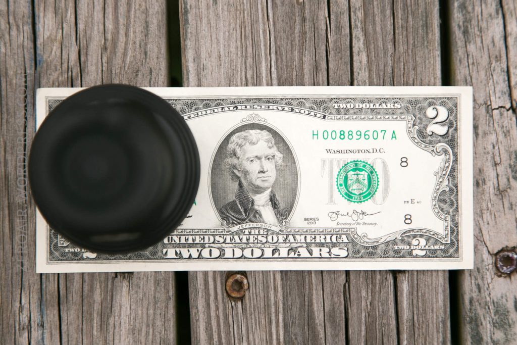 A two-dollar bill is placed on a weathered wooden surface with a black round object partially covering the left side of the bill.