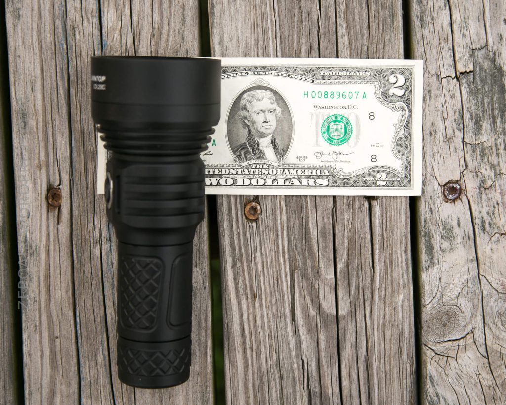 A black flashlight is placed next to a U.S. two-dollar bill on a weathered wooden surface, showing the relative size comparison between the flashlight and the bill.
