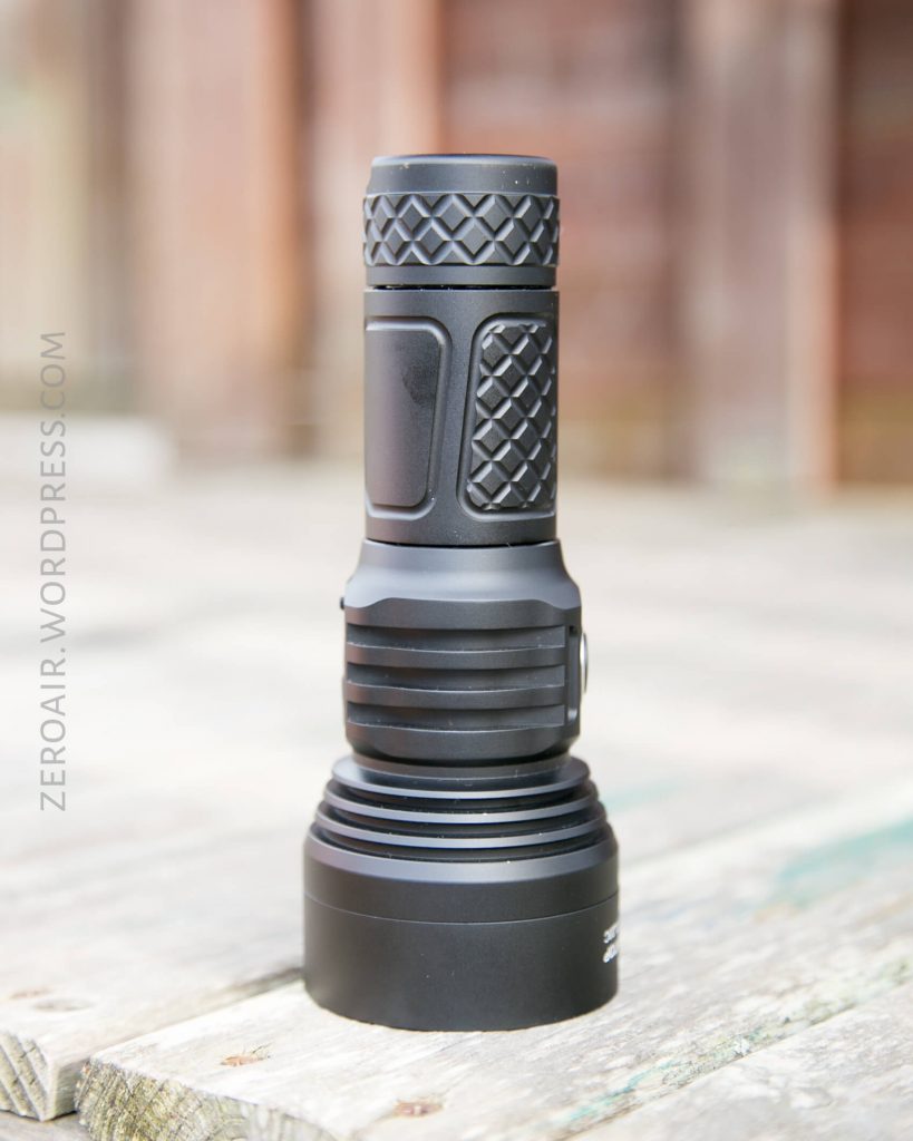 A black handheld flashlight with a textured grip stands upright on a weathered wooden surface. The background is blurred.