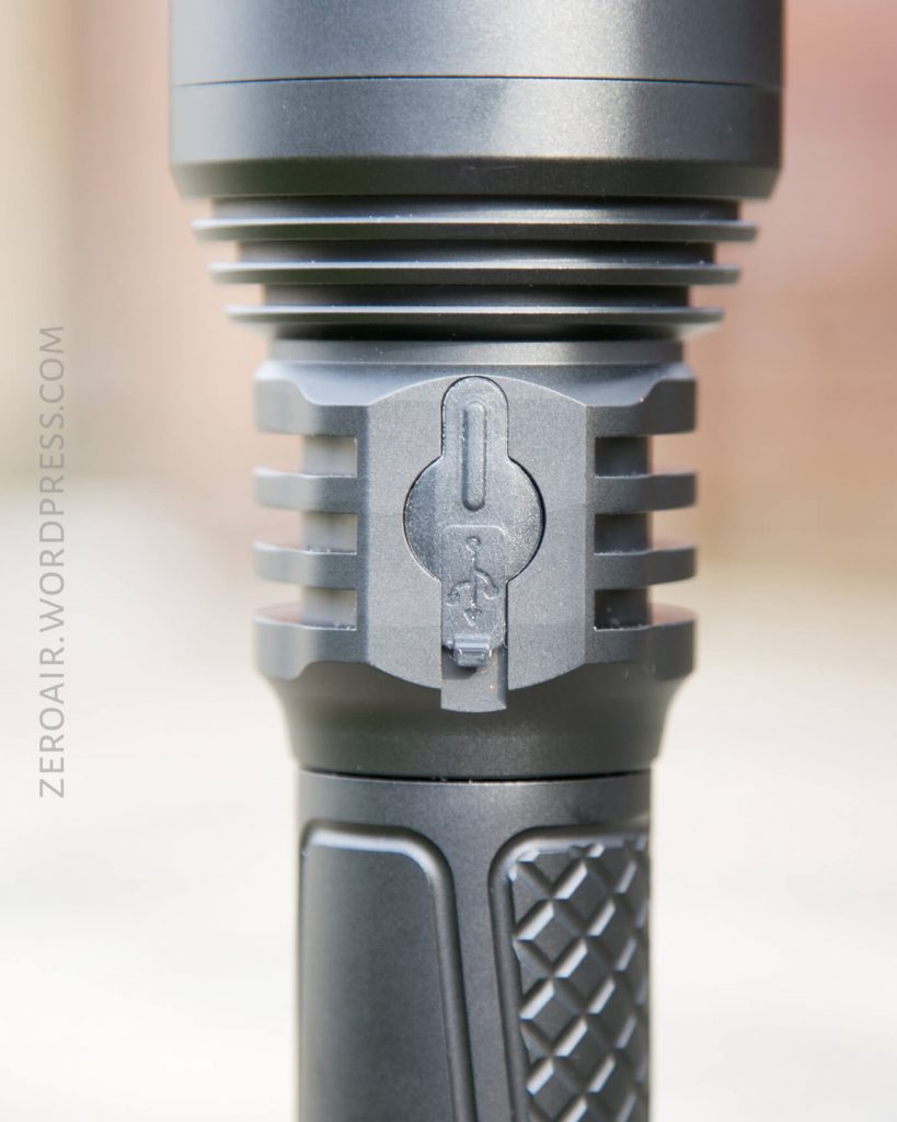 Close-up of a black flashlight showing detailed grooves, a textured grip, and a rubber cover labeled with a USB symbol over a charging port. The words ZEROAIR.WORDPRESS.COM are visible along the left side.