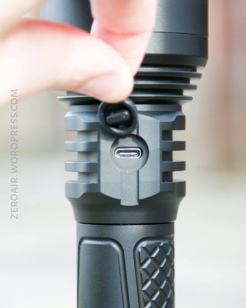 A close-up of a hand opening a rubber cover to reveal a USB-C charging port on a black flashlight. The flashlight has a textured grip and vertical ridges above the charging port.