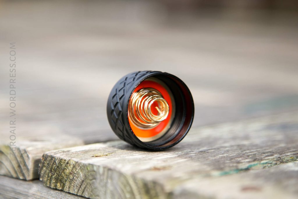 A close-up of a black, textured flashlight tailcap with a visible metal spring inside, resting on a weathered wooden surface. The background is blurred.
