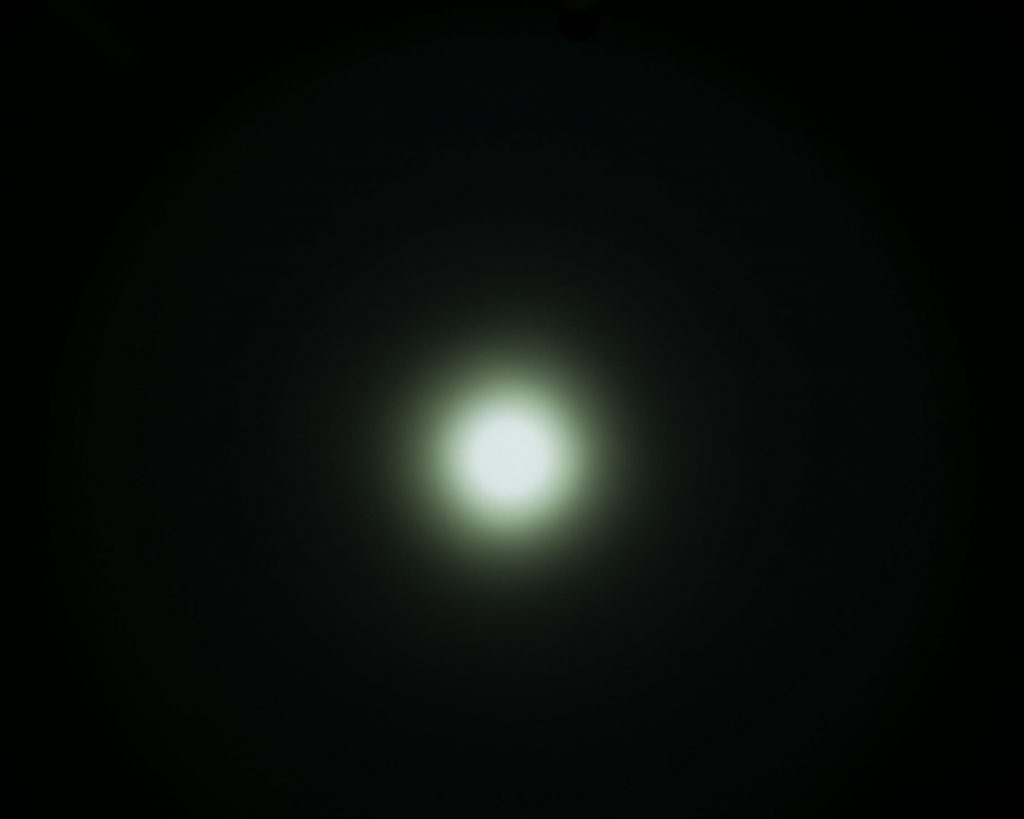 A bright white circular light is centered against a dark, almost black background, with a soft, diffuse glow radiating outward. The surrounding edges gradually fade from light to dark.