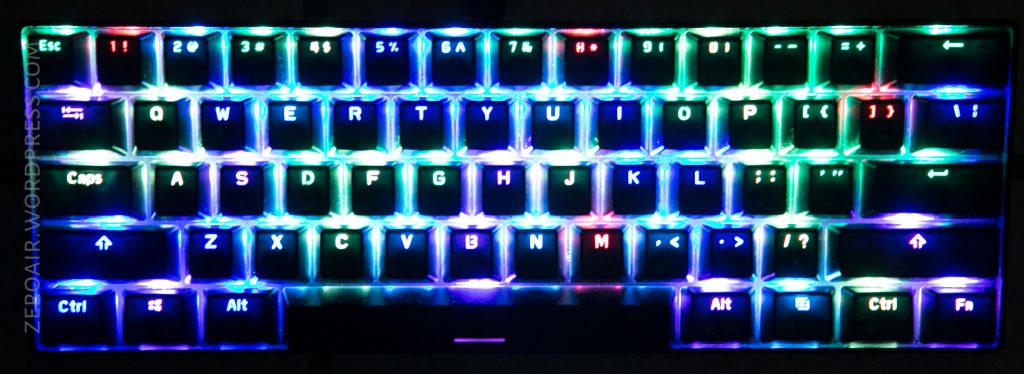 A compact keyboard with RGB backlighting displays a rainbow of colors across the keys, including blue, green, and red hues. Keys are clearly visible with bright, colorful illumination.
