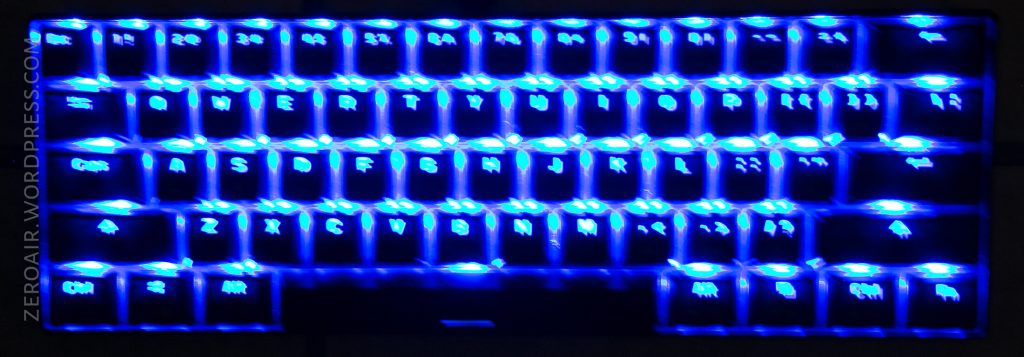 A close-up of a compact keyboard with bright blue LED backlighting, showing both English and another languages characters on the keys. The keys are illuminated, and the image is slightly blurred.