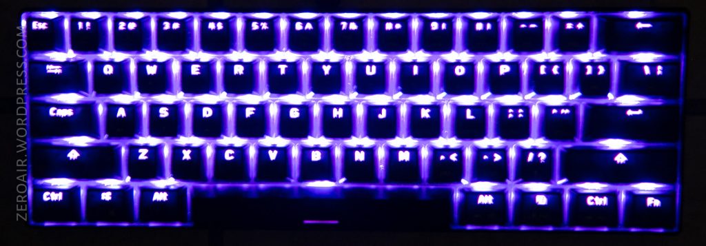A compact mechanical keyboard with its keys illuminated by bright purple LED backlighting, viewed from above in a dark setting. The key legends are visible, and the left side has “zeroair.wordpress.com” faintly displayed.