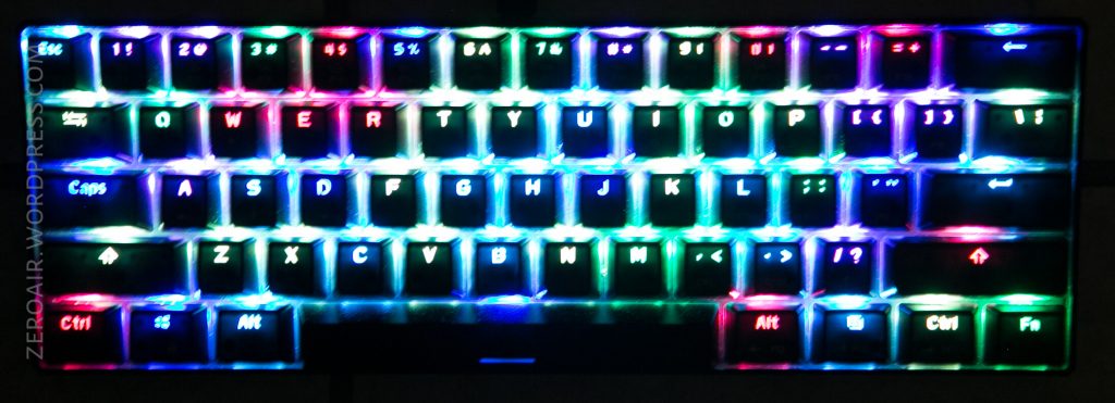 A compact mechanical keyboard with keys illuminated by multicolored RGB backlighting, arranged in horizontal lines of red, green, and blue hues. The keyboard has a standard QWERTY layout.