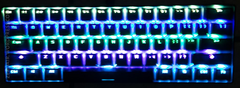 A compact keyboard is illuminated with green, aqua, and purple backlighting. The keys are arranged in a grid layout with transparent keycaps, and the letters and numbers are visible.