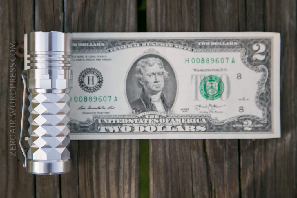 A small metallic flashlight with a textured grip is placed next to a U.S. two-dollar bill on a wooden surface. The flashlight and bill are aligned side by side for size comparison.