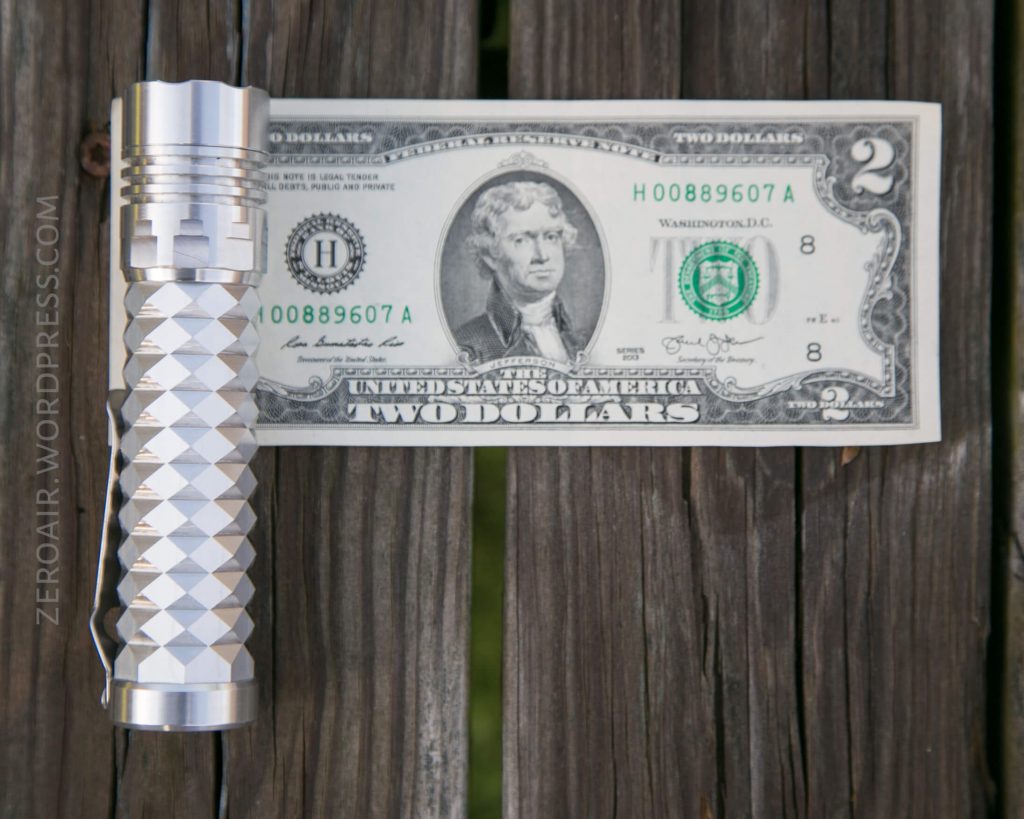 A silver flashlight with a textured grip lying next to a two-dollar bill on a wooden surface. The flashlight is about the same length as the bill.