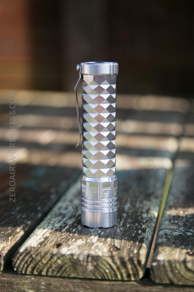 A metallic flashlight with a textured, diamond-patterned handle stands upright on a weathered wooden surface outdoors. The background is out of focus.