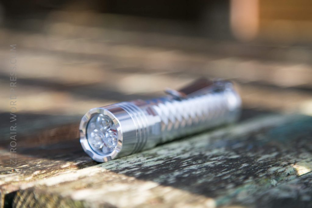 A metallic flashlight lying on a weathered wooden surface, with the lens facing the camera and the background out of focus.