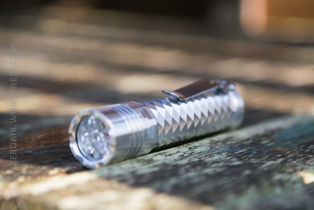 A metallic flashlight with a textured, diamond-patterned body rests on a weathered wooden surface. The flashlight has a pocket clip attached.