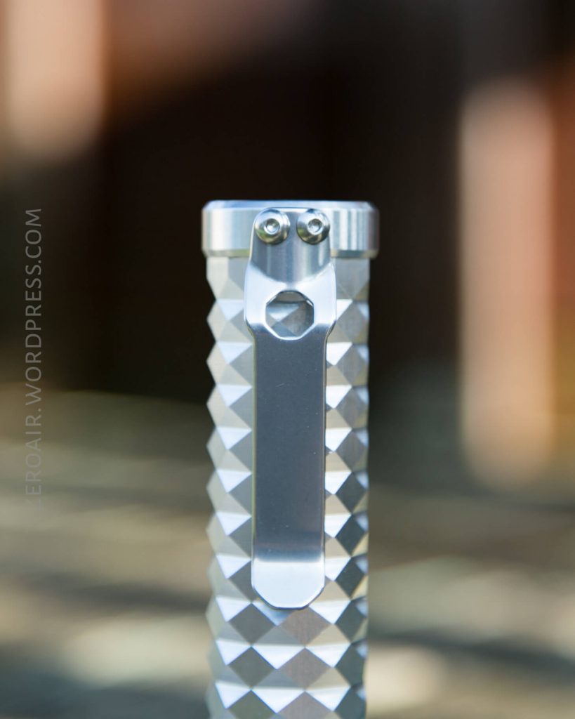 Close-up of a silver, diamond-patterned metal object with a pocket clip and two visible screws at the top. Background is blurred. The text EROLFAR.WORDPRESS.COM is written vertically on the left side.