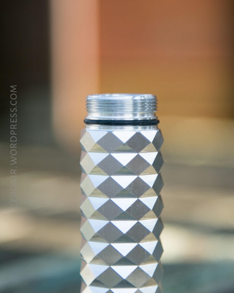 A close-up of a metallic, diamond-patterned cylindrical object with threaded top, possibly part of a tool or container, standing vertically with a blurred background. ZERO4R.WORDPRESS.COM is visible on the left side.