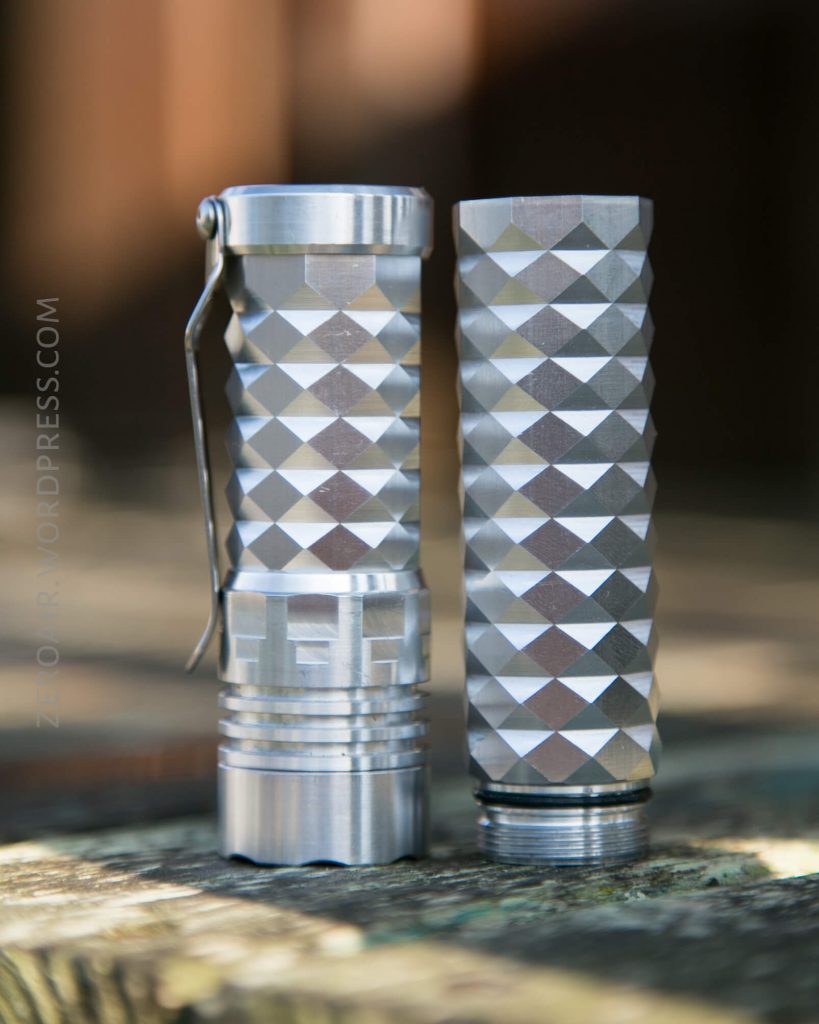Two cylindrical, silver metal flashlights with a diamond-patterned grip are shown standing upright on a wooden surface. One flashlight is assembled while the other has its end cap removed, revealing the threads.