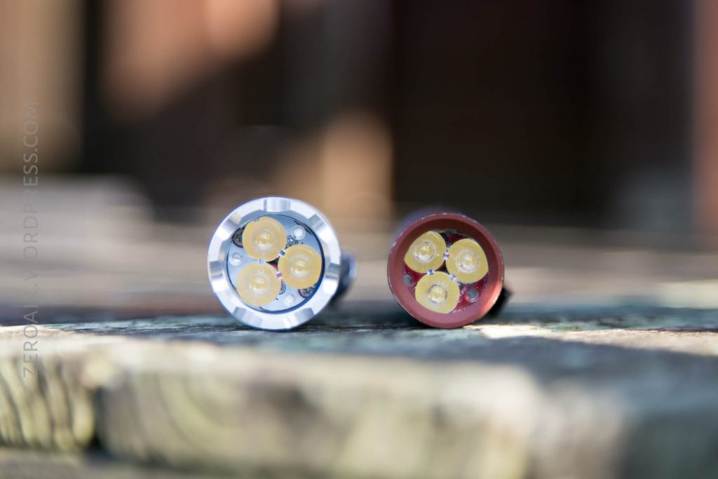 Two small, round flashlight heads with four yellow LED bulbs each are positioned side by side on a wooden surface. The left flashlight has a silver casing; the right one has a red casing. The background is blurred.