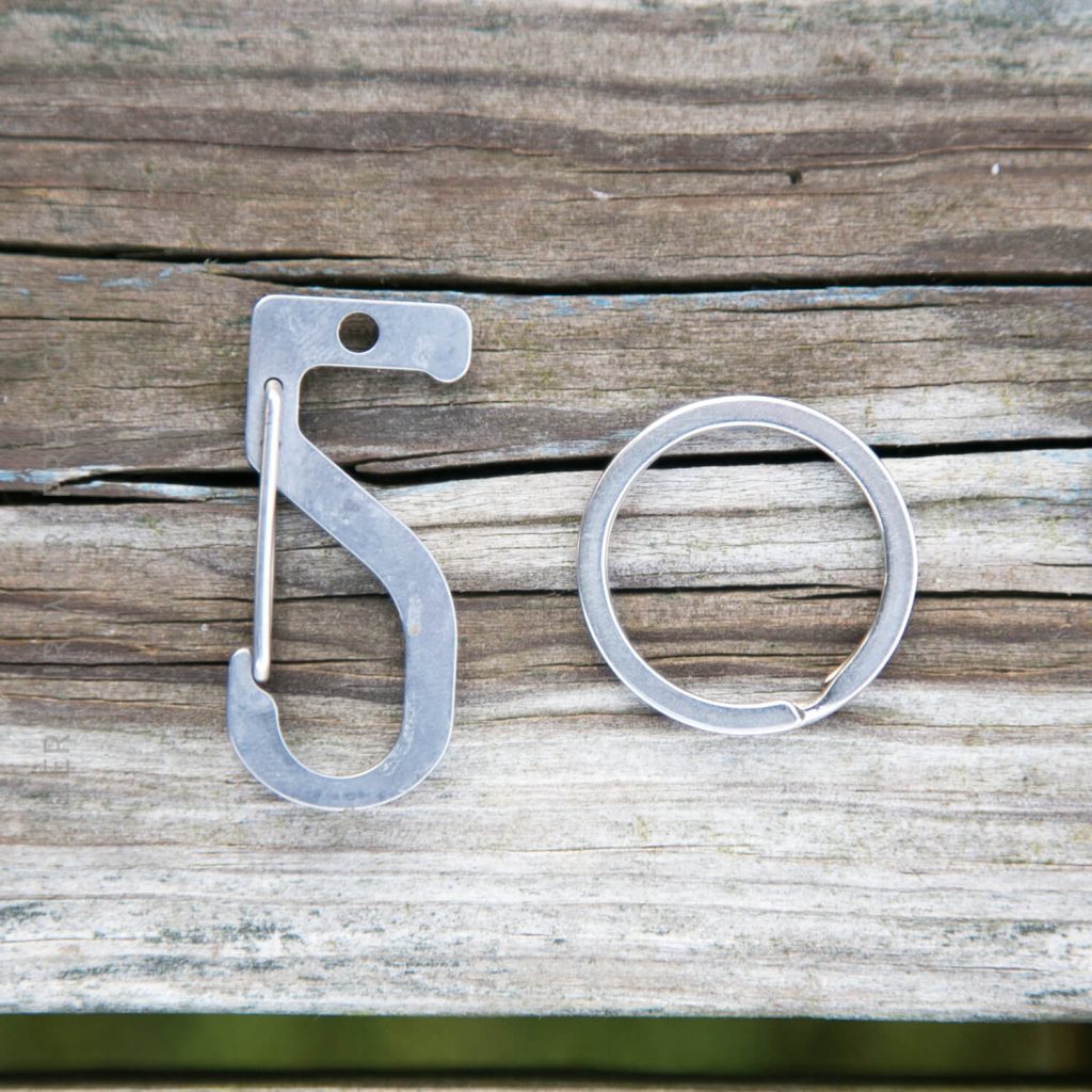 A silver S-shaped carabiner and a round metal key ring are placed side by side on weathered wooden boards.