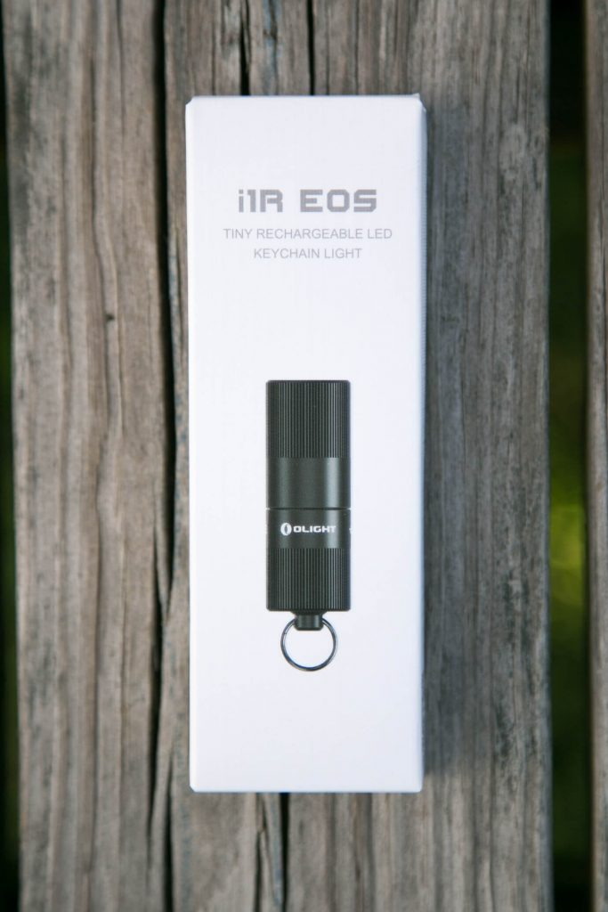 A small white box with “i1R EOS Tiny Rechargeable LED Keychain Light” printed on it, showing an image of a black cylindrical keychain flashlight, placed on weathered wooden planks.