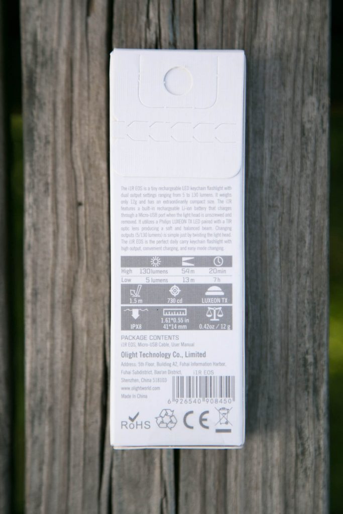 A white product box for an Olight keychain flashlight stands upright on weathered wooden boards. The back label shows product information, technical details, specifications, and a barcode.