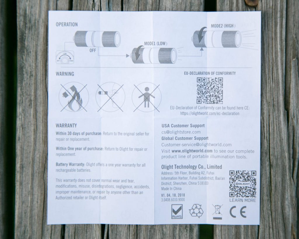 Instruction sheet for a flashlight on a wooden surface, showing diagrams for operation, warnings, warranty, battery info, customer support contacts, QR codes, and compliance details. Text and graphics are printed in black on white paper.