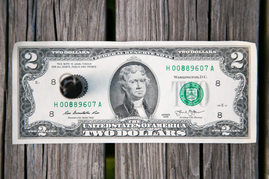 A US two-dollar bill with a hole punched through its left side is placed on a weathered wooden surface. The bill features the portrait of Thomas Jefferson.