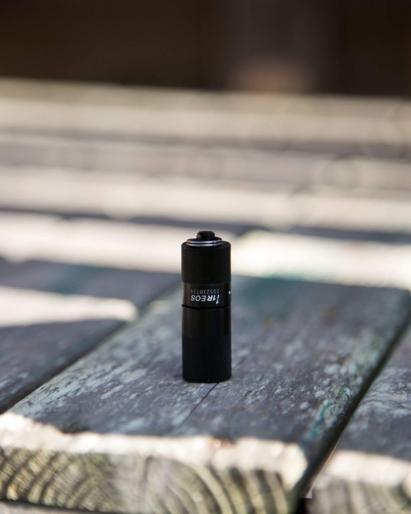 A small black cylindrical object labeled Nitecore standing upright on a weathered wooden surface with blurred horizontal planks in the background.
