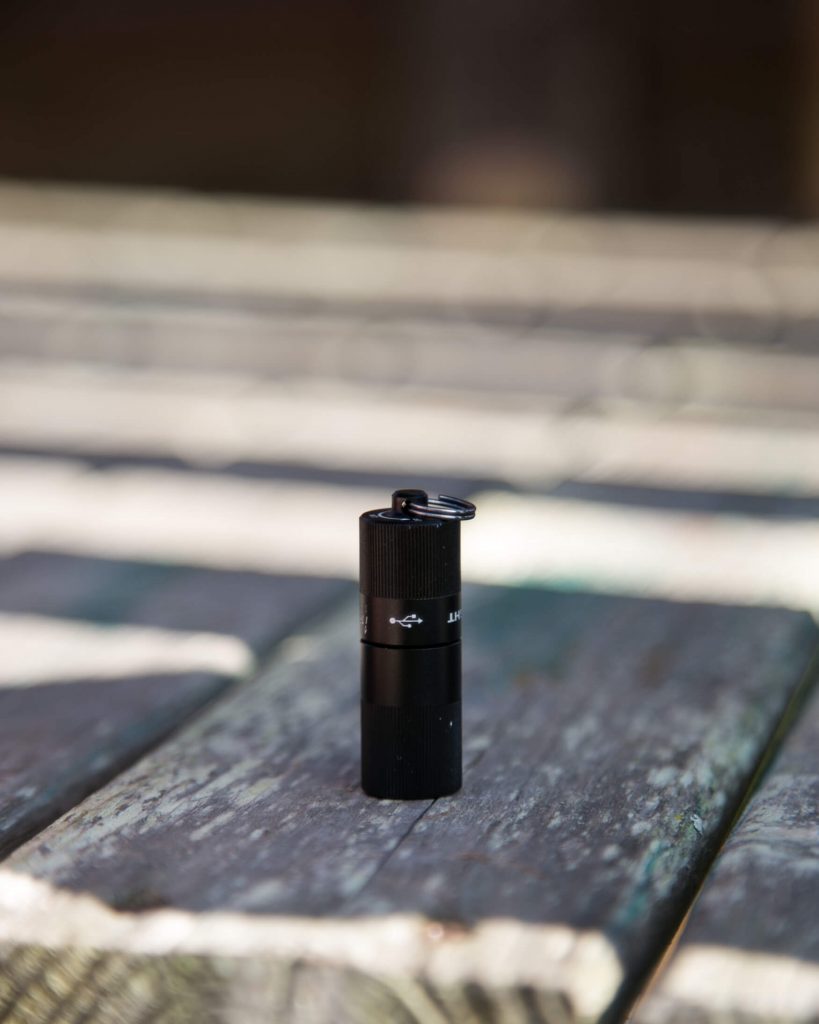 A small black flashlight with a keyring attachment stands upright on a weathered wooden surface with sunlight casting shadows across the planks.