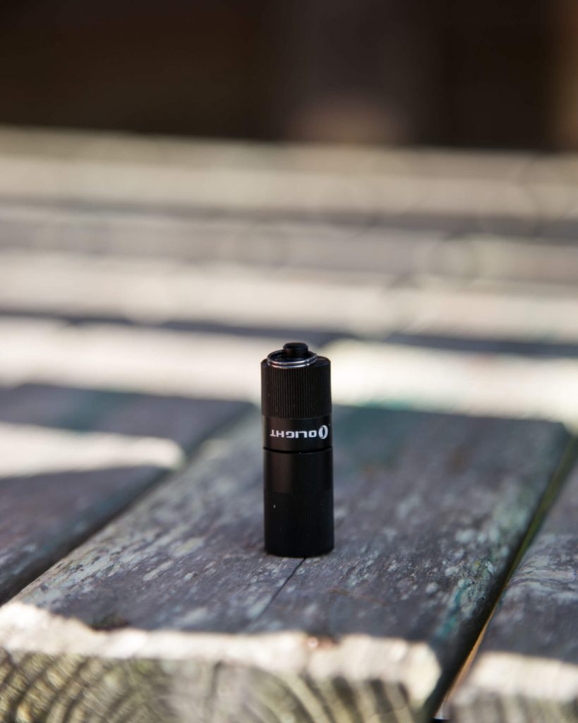 A small black Olight flashlight stands upright on a weathered wooden surface with blurred outdoor surroundings in the background.