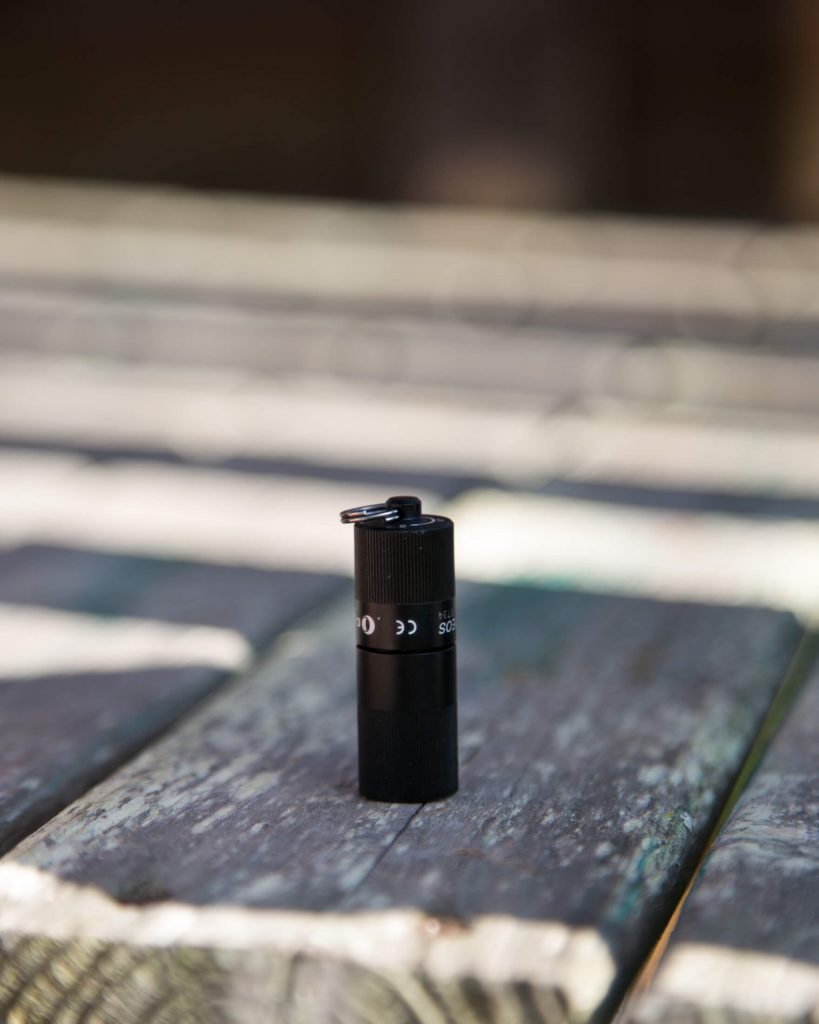 A small black cylindrical object with a keychain loop on top is standing upright on a weathered wooden surface with sunlight and shadows.