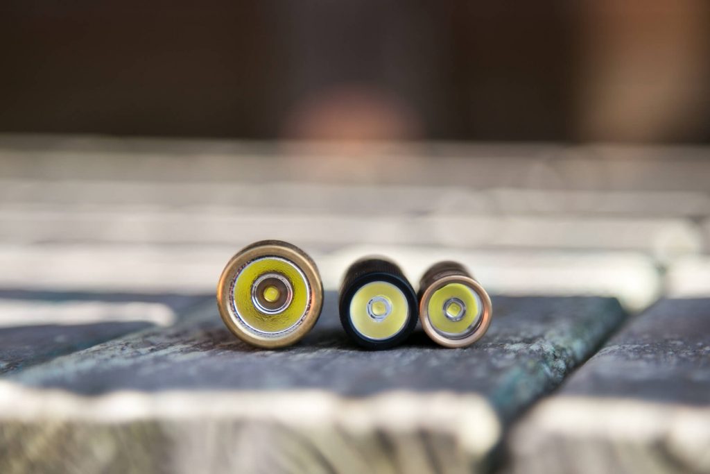 Three batteries of different sizes are lying horizontally on a wooden surface, viewed from the positive terminal end. The focus is on the batteries, while the background is blurred.