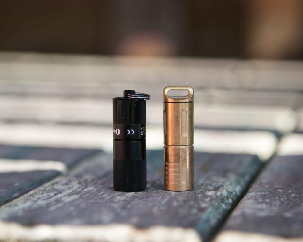 A small black flashlight and a small brass flashlight are standing upright side by side on a wooden surface with a blurred background.