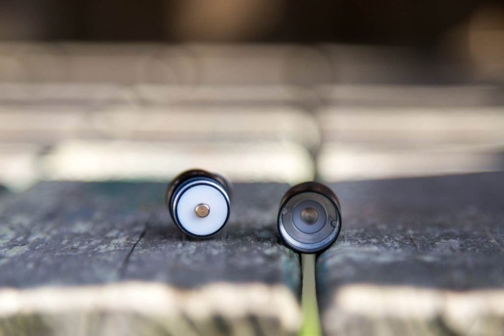 Two small cylindrical metal objects lie parallel on a weathered wooden surface, both with one end facing the camera. The left cylinder shows a white circular center, while the right one has a dark, hollow opening.