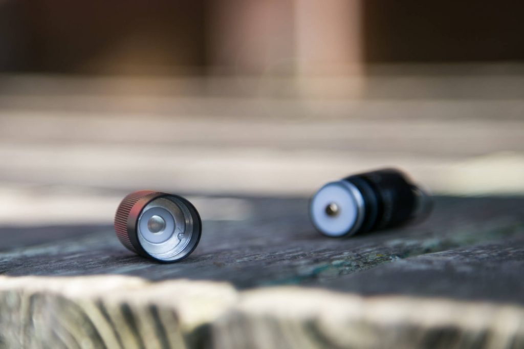 Two small cylindrical objects with metal ends, possibly batteries or flashlight components, are lying on a rough wooden surface with a blurred background.