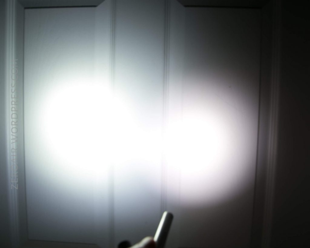 A white door with vertical panels is illuminated by two overlapping circles of bright light. A hand holding a cylindrical object, possibly a flashlight, appears at the bottom of the image.