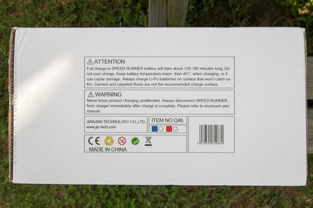 A white product box viewed from above displays printed safety warnings, manufacturer information, certification icons, a barcode, and the item number Q46. The box is resting on grass near a wooden post.