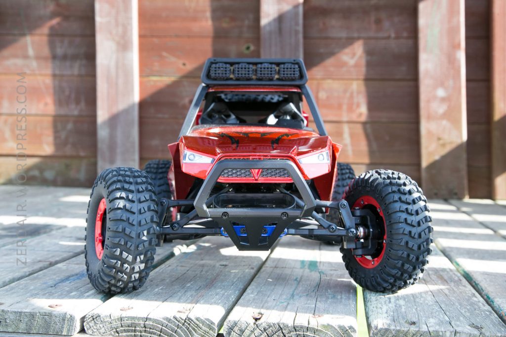 A red remote-controlled off-road car with large, rugged tires is positioned on a wooden surface outdoors, with a wooden fence in the background.
