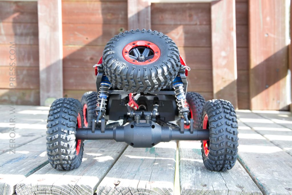 A close-up, rear view of a remote-controlled off-road vehicle with large, knobby tires, red wheel rims, and a mounted spare tire, sitting on a wooden surface with a wooden fence in the background.