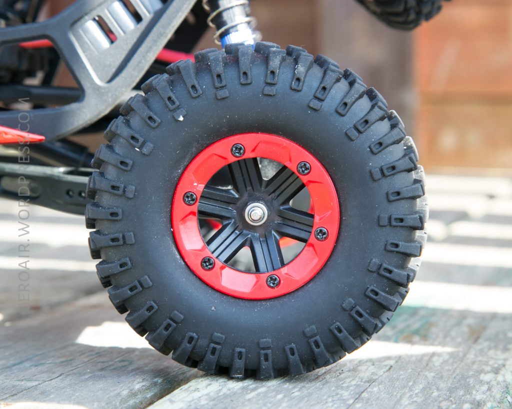 Close-up of a black rubber tire with a red and black rim attached to a toy vehicle, positioned on a wooden surface. The tire has deep treads and visible screws around the rim.