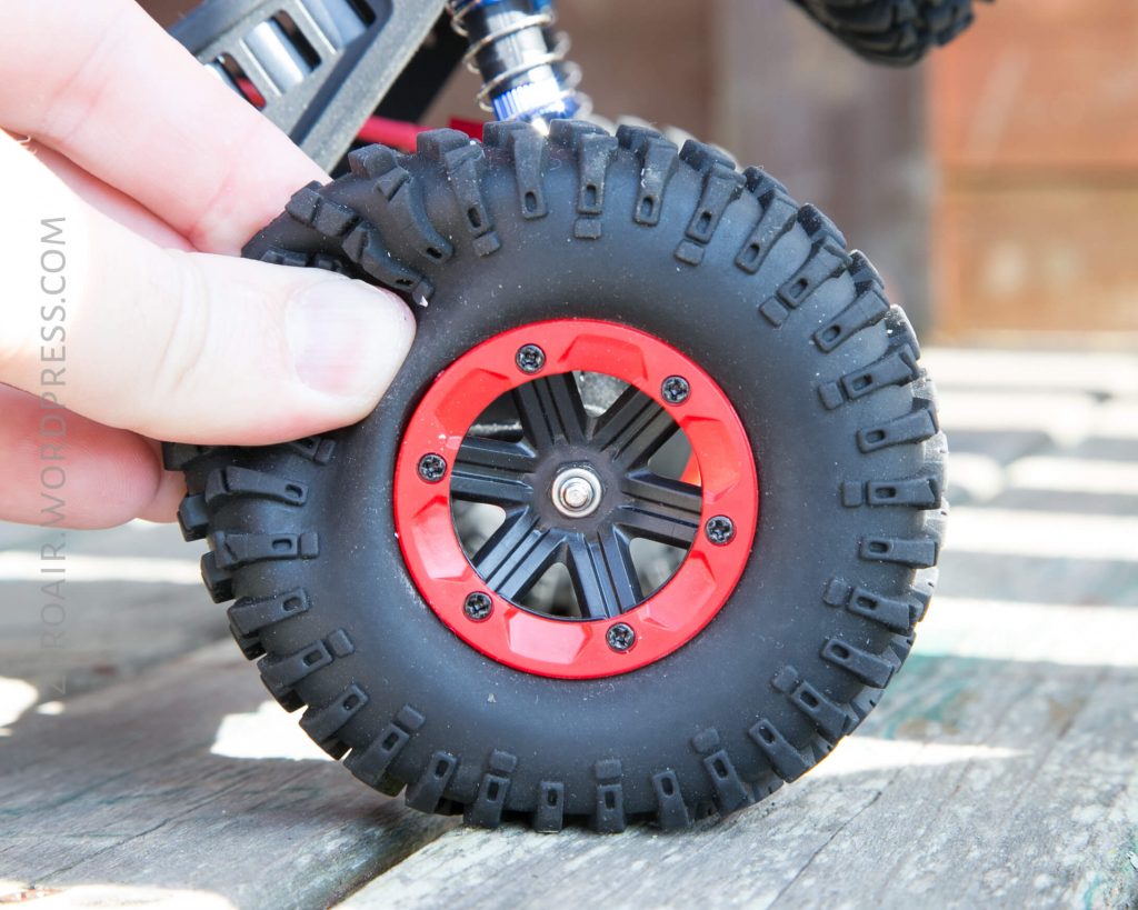 A close-up of a persons hand holding the tire of a remote-controlled vehicle with a black rubber tire and red rim, placed on a wooden surface.