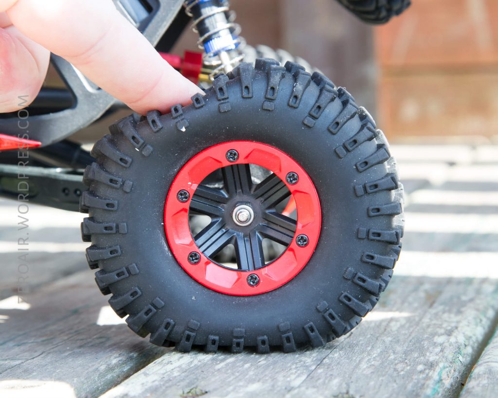 A close-up of a person’s finger pressing on the top of a black, rugged tire with a red and black rim, attached to a vehicle, possibly a remote-controlled car. The background is slightly blurred.